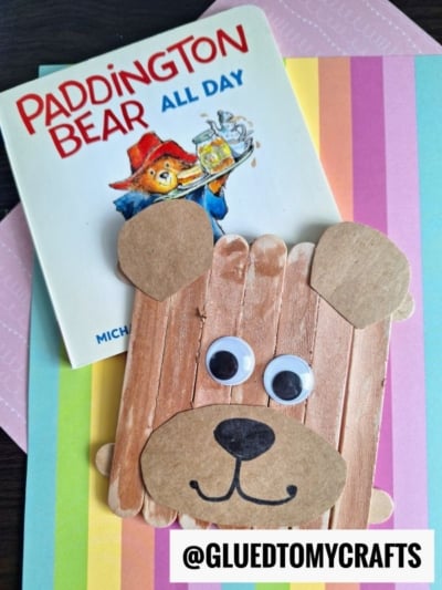 A Paddington Bear All Day book sits on pastel striped paper, next to a bear face craft made from popsicle sticks, googly eyes, and brown paper—perfect for summer craft ideas. @GLUEDTOMYCRAFTS is written at the bottom.