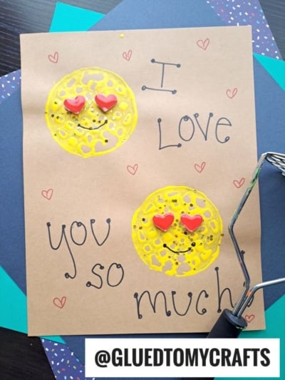 A handmade card featuring two yellow smiley faces with red heart-shaped eyes, painted on brown paper using a potato masher stamped craft technique. The card reads "I love you so much" and is adorned with small red hearts, with a craft roller nearby.