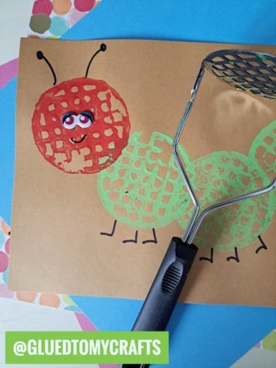 A childrens potato masher stamped craft of a caterpillar made with painted prints on brown paper, featuring a smiling face with eyes and antennae. The image includes a potato masher and colorful papers. @GLUEDTOMYCRAFTS is labeled below.