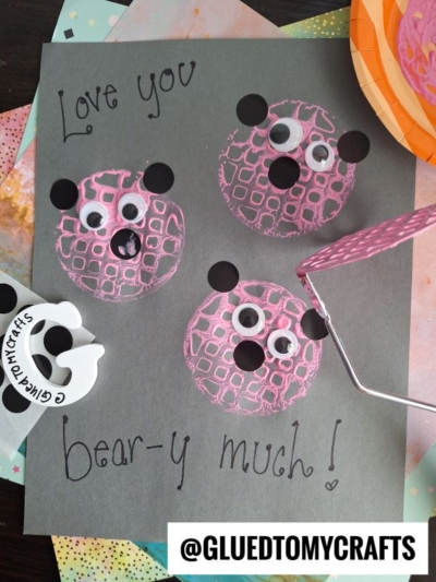 A craft project shows three pink bear faces made from paint, googly eyes, and black paper ears on black paper. Text reads Love you bear-y much! and a handle with pink tape is visible. @GLUEDTOMYCRAFTS is at the bottom.
