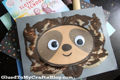 A handmade sloth face craft with large googly eyes and a smile, created using brown and cream paint and paper, sits on a gray background. A children’s book about sloths is partially visible behind the craft.