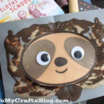 A handmade sloth face craft with large googly eyes and a smile, created using brown and cream paint and paper, sits on a gray background. A children’s book about sloths is partially visible behind the craft.