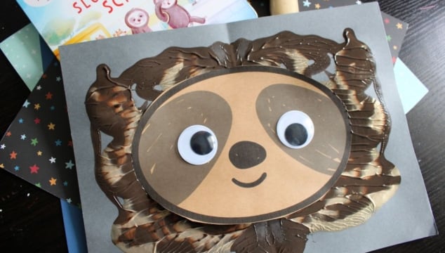 A handmade sloth face craft with large googly eyes and a smile, created using brown and cream paint and paper, sits on a gray background. A children’s book about sloths is partially visible behind the craft.
