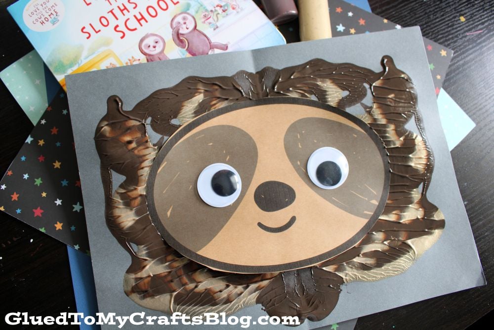 A handmade sloth face craft with large googly eyes and a smile, created using brown and cream paint and paper, sits on a gray background. A children’s book about sloths is partially visible behind the craft.