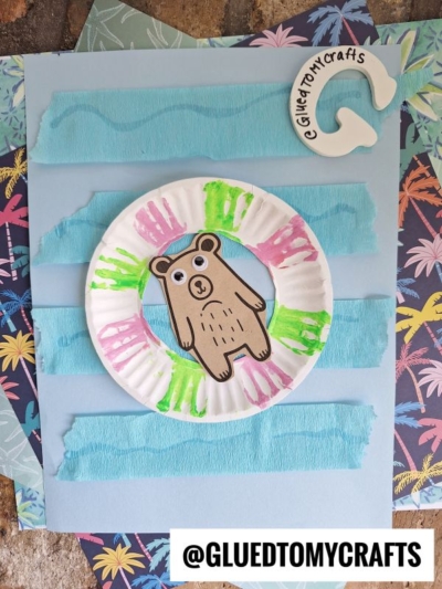 A paper plate lazy river craft for kids, featuring a green and pink floatation ring with a cartoon bear at the center, set on blue paper strips and surrounded by tropical-themed paper for a fun summer scene.