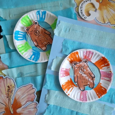 Two paper plate bear crafts with colorful painted borders are displayed on blue paper, decorated with horizontal strips and hibiscus flower cutouts. The Instagram handle @GLUEDTOMYCRAFTS is shown at the bottom.
