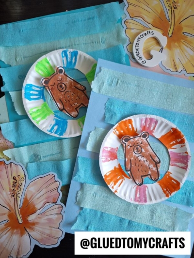 Two paper plate bear crafts with colorful painted borders are displayed on blue paper, decorated with horizontal strips and hibiscus flower cutouts. The Instagram handle @GLUEDTOMYCRAFTS is shown at the bottom.