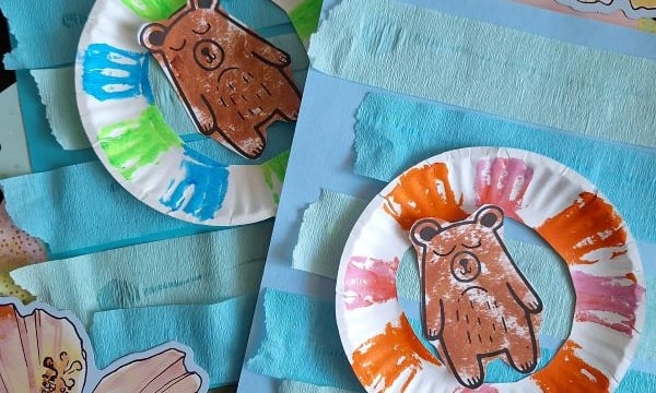 Two paper plate bear crafts with colorful painted borders are displayed on blue paper, decorated with horizontal strips and hibiscus flower cutouts. The Instagram handle @GLUEDTOMYCRAFTS is shown at the bottom.