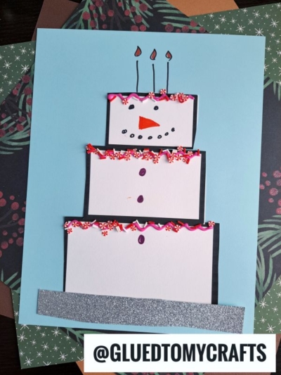 A paper craft displays a charming snowman cake as a three-tiered dessert, decorated with pink icing, sprinkles, and candles. The snowman features a carrot nose, eyes, mouth, and buttons drawn in marker on a blue background.
