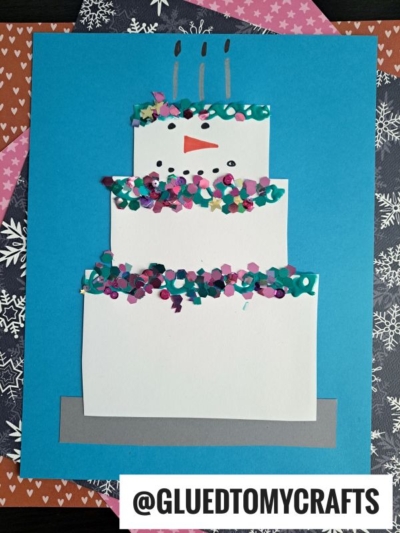 A handmade snowman cake craft on blue paper, featuring a cheerful snowman face with an orange nose, colorful confetti, and three candles. The username @GLUEDTOMYCRAFTS appears at the bottom. Perfect for a snowman cake themed project!.