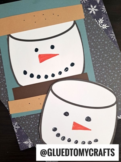 Two handmade paper snowman faces, resembling s'mores with orange carrot noses, black dotted eyes and smiles, plus brown hat shapes. Displayed on a snowy-patterned surface. @GLUEDTOMYCRAFTS is written at the bottom.