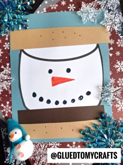 A handmade paper snowman face craft made with colored paper strips, black marker, and an orange nose. Decorative snowflakes and a cute paper snowman s'mores figurine are nearby, with a @GLUEDTOMYCRAFTS watermark at the bottom.