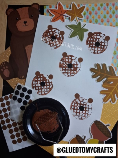 Children’s fall craft with brown bear cutouts, leaf and acorn shapes, and a white paper featuring potato masher stamped bear faces made with brown paint and black dot stickers. Paint and stickers are placed nearby.
