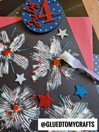 A Fourth of July craft features painted firework bursts on black paper, glittery red, silver, and blue stars, and a “July 4” sign. A toothbrush with white paint is also visible. The handle @GLUEDTOMYCRAFTS is at the bottom.