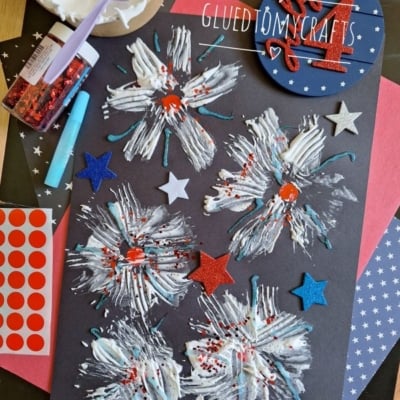 A black paper displays white and blue painted firework designs with red glitter and star stickers, surrounded by craft supplies like red dot stickers, glitter, a palette of white paint, and a @GLUEDTOMYCRAFTS label.