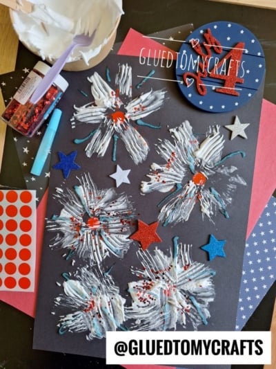 A black paper displays white and blue painted firework designs with red glitter and star stickers, surrounded by craft supplies like red dot stickers, glitter, a palette of white paint, and a @GLUEDTOMYCRAFTS label.