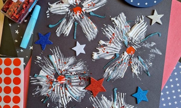 A black paper displays white and blue painted firework designs with red glitter and star stickers, surrounded by craft supplies like red dot stickers, glitter, a palette of white paint, and a @GLUEDTOMYCRAFTS label.