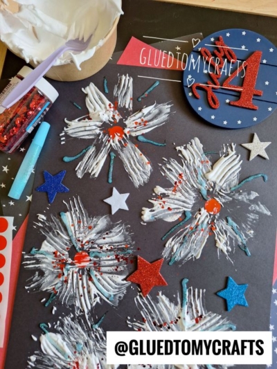 A black paper features white paint fireworks with red centers and red glitter, surrounded by blue and red glitter stars. Craft supplies, including glue, foam brush, and glitter, are visible beside the artwork. @GLUEDTOMYCRAFTS text is at the bottom.