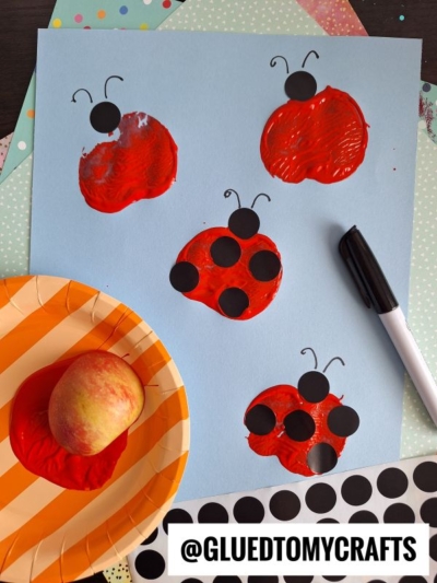 A blue paper displays an APPLE STAMPED CRAFT: ladybugs made from red apple prints, with drawn legs and antennae, plus black sticker dots for spots. An apple, a plate with red paint, a black marker, and stickers are nearby.