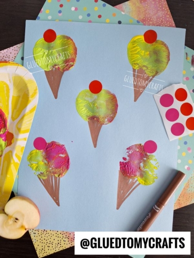 Blue paper displays five apple stamped craft ice cream cones with red dot cherries, made using apple halves and paint. A brown marker and patterned papers lie nearby. The handle @GLUEDTOMYCRAFTS is printed at the bottom.