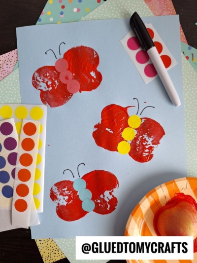 A blue paper features an APPLE STAMPED CRAFT with three red butterfly prints made from apple halves, each decorated with colored dot stickers for the body and black marker antennae. Supplies like stickers, marker, and an apple are nearby.