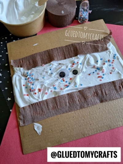 A square brown paper features rows of brown crepe paper and white shaving cream with red and blue star confetti and two googly eyes. @GLUEDTOMYCRAFTS is labeled on the image. Craft supplies are visible nearby.