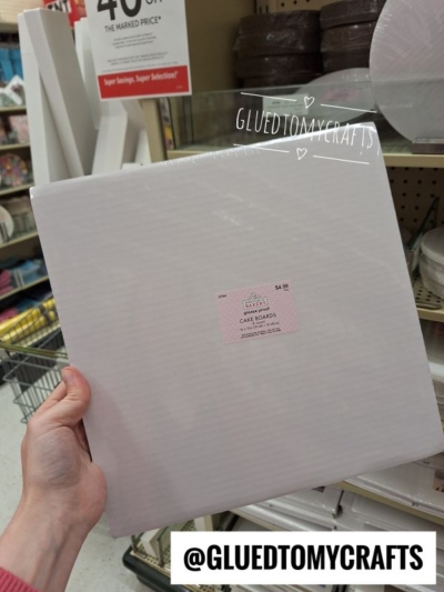 A hand holds a pack of 12-inch square white cake boards in a store aisle. The product has a pink label, and there is a @GLUEDTOMYCRAFTS watermark on the image. Craft supplies are visible in the background.