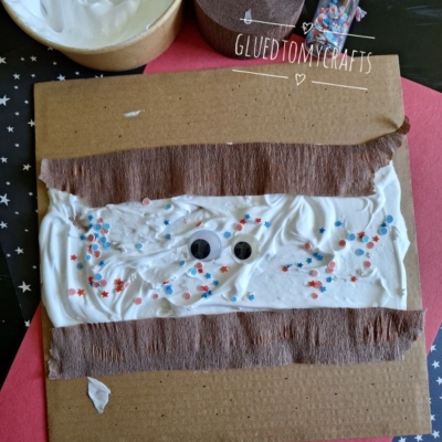 A craft project: brown cardboard with white glue or cream spread in the center, googly eyes, and red and blue star-shaped confetti, with brown tissue strips and a “@GLUEDTOMYCRAFTS” label.