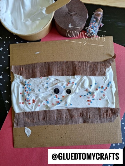 A craft project: brown cardboard with white glue or cream spread in the center, googly eyes, and red and blue star-shaped confetti, with brown tissue strips and a “@GLUEDTOMYCRAFTS” label.
