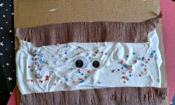 A craft project: brown cardboard with white glue or cream spread in the center, googly eyes, and red and blue star-shaped confetti, with brown tissue strips and a “@GLUEDTOMYCRAFTS” label.