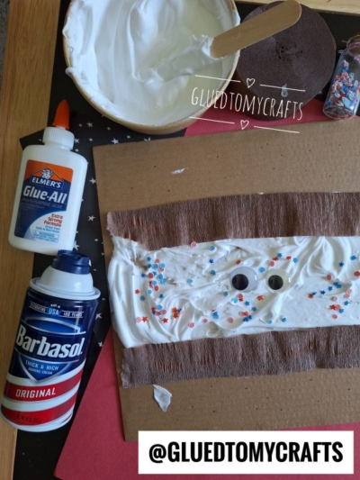 A craft project in progress: a piece of cardboard with white shaving cream, colorful star-shaped sprinkles, two googly eyes, and strips of brown paper. Nearby are glue, shaving cream, a bowl of white mixture, and craft supplies.