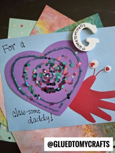 A handmade HANDPRINT FATHER'S DAY card features a purple heart with sequins and glitter, layered on blue paper with a red handprint crab. Text reads “For a Claw-some daddy!” and “@GLUETOMYCRAFTS” is at the bottom.