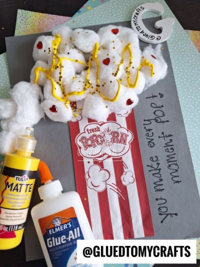 A handmade cotton ball Valentine's Day popcorn craft features cotton balls as popcorn, yellow paint as butter, and red hearts on a paper popcorn box. The gray background displays “You make every moment pop!” with visible craft supplies.
