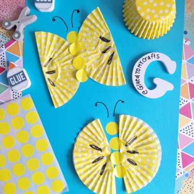 Yellow polka dot cupcake liners are crafted into butterflies and a caterpillar on a blue paper background, with craft supplies like glue, scissors, and patterned papers nearby. @GLUETOMYCRAFTS is labeled at the bottom.