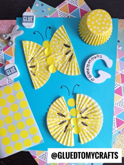 Yellow polka dot cupcake liners are crafted into butterflies and a caterpillar on a blue paper background, with craft supplies like glue, scissors, and patterned papers nearby. @GLUETOMYCRAFTS is labeled at the bottom.