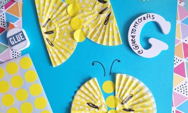 Yellow polka dot cupcake liners are crafted into butterflies and a caterpillar on a blue paper background, with craft supplies like glue, scissors, and patterned papers nearby. @GLUETOMYCRAFTS is labeled at the bottom.