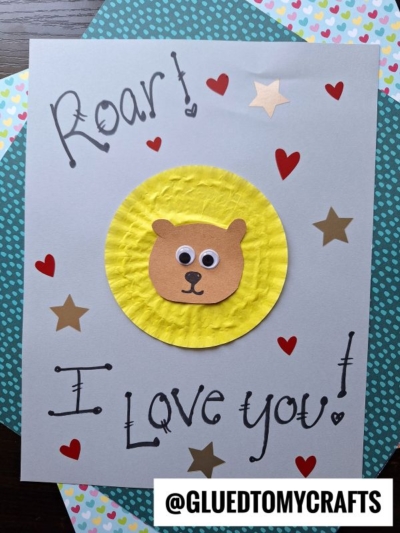 A handmade cupcake liner card craft featuring a bear face on a yellow circle, surrounded by red hearts and gold stars. “Roar! I Love You!” is written on the card, with the handle @GLUETOMYCRAFTS at the bottom.