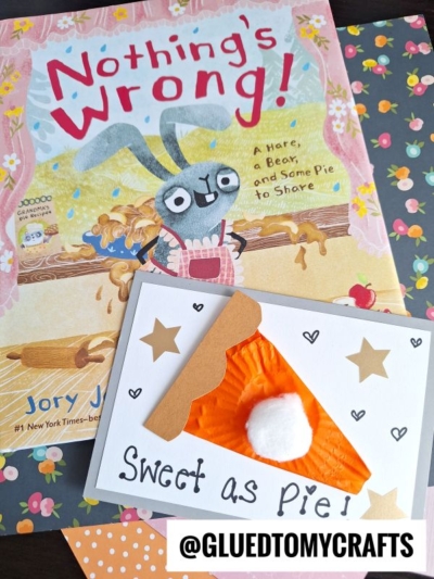 A children’s book titled Nothing’s Wrong! with a bunny on the cover sits beside a handmade cupcake liner card shaped like pie, topped with a white pom-pom and “Sweet as Pie!” written below, all on patterned paper.