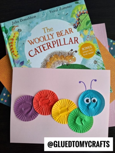 A colorful caterpillar craft made from cupcake liners and googly eyes is on pink paper, with The Woolly Bear Caterpillar book and patterned papers in the background—a fun example of INSECT CRAFT IDEAS. @GLUETOMYCRAFTS is written at the bottom.