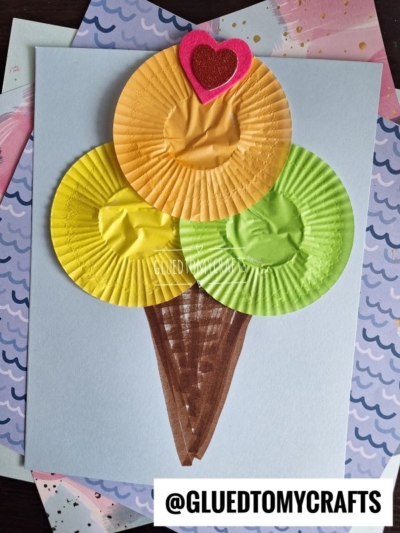 A craft ice cream cone made with brown marker and three colored cupcake liners (orange, yellow-green, and yellow), topped with a pink foam heart on light blue paper—perfect inspiration for cupcake liner card crafts. The image includes the text @GLUEDTOMYCRAFTS.