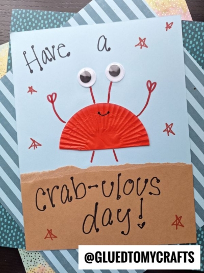 This cupcake liner card crafts a charming red crab with googly eyes on blue paper, surrounded by stars. The text reads “Have a crab-ulous day!” with @GLUEDTOMYCRAFTS at the bottom.