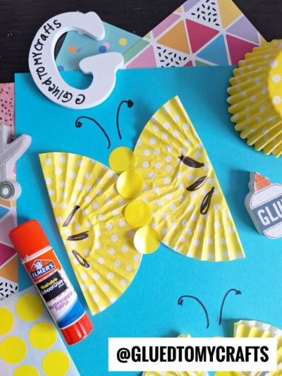 A yellow polka-dot butterfly made from cupcake liners is glued to blue paper. A glue stick, yellow stickers, a paper G, and other craft supplies are arranged around the craft. Text reads @GLUEDTOMYCRAFTS.