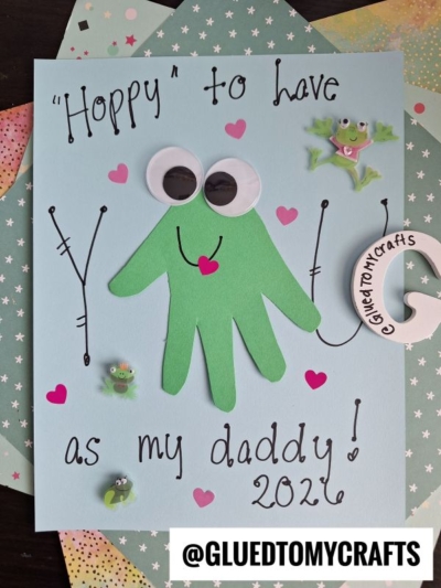 A handmade HANDPRINT FATHER'S DAY card features a green handprint frog with googly eyes and a pink heart on blue paper. It reads, Hoppy to have you as my daddy! 2024, and is decorated with frog stickers and colorful paper.