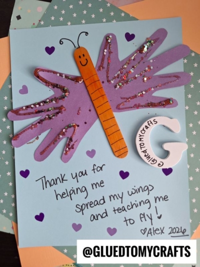 A handmade HANDPRINT FATHER'S DAY card featuring a purple butterfly crafted from glittery handprints and a craft stick. A heartfelt note reads: Thank you for helping me spread my wings and teaching me to fly! Alex 2026, with heart decorations.