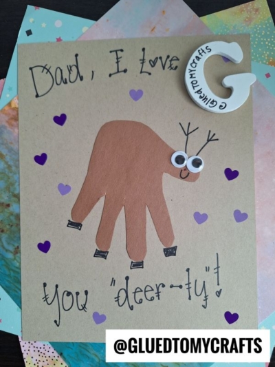 A HANDPRINT FATHER'S DAY card features a brown handprint shaped like a deer with googly eyes, antlers, and a smile, surrounded by purple hearts. The text reads “Dad, I love you ‘deer-ly’!”.