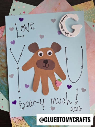 A handmade HANDPRINT FATHER'S DAY card featuring a bear crafted from a brown handprint, googly eyes, and a pink heart. The message reads Love you bear-y much! 2026. Decorated with hearts and colorful papers all around.