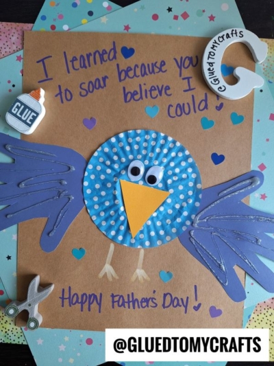 A handmade HANDPRINT FATHER'S DAY card features a blue bird crafted from paper and a blue polka-dot cupcake liner, with a hand-written message saying, I learned to soar because you believed I could. Craft supplies and decorations surround the card.