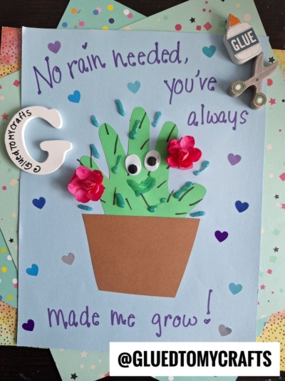 A handmade card with a green paper cactus in a brown pot, decorated with googly eyes, pink flowers, and blue raindrops. Perfect for a HANDPRINT FATHER'S DAY gift, it features purple text: No rain needed, you’ve always made me grow!.