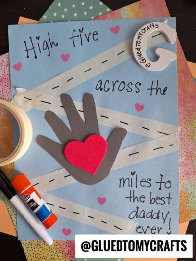 A HANDPRINT FATHER'S DAY card features a paper hand and red heart on blue, with roads and the message: High five across the miles to the best daddy ever! Craft supplies and patterned paper surround this heartfelt creation.