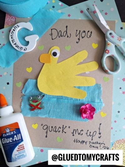 A handmade HANDPRINT FATHER'S DAY card reads “Dad, you ‘quack’ me up!” with a yellow handprint duck, a googly eye, blue fabric, a small frog sticker, flower, glue, scissors, and heart stickers on a colorful background.